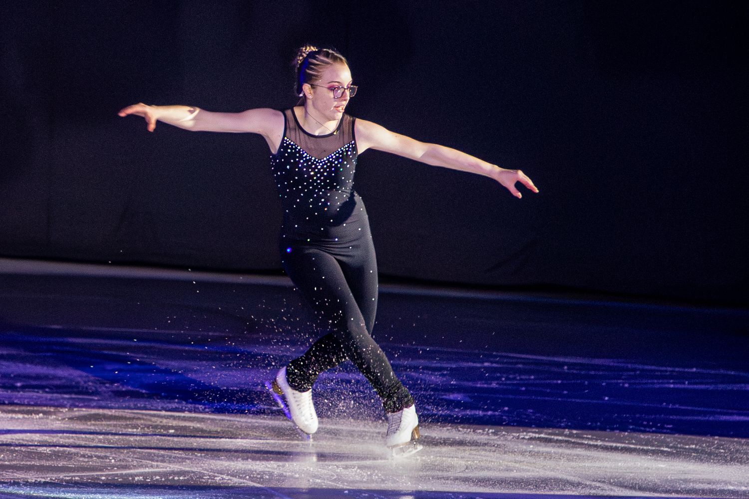 Female figure skater in black outfit gracefully performing on ice rink with arms extended.