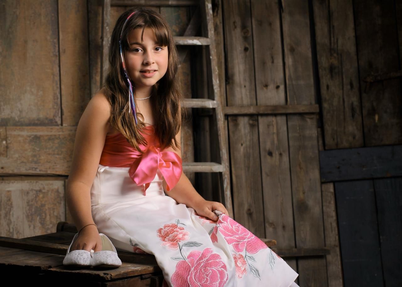 A person in a white dress with pink floral accents poses against a rustic wooden backdrop in a portrait session.