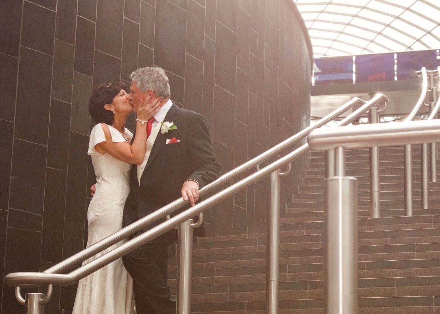 Bride and groom embrace on modern stairs with dark brick wall backdrop.