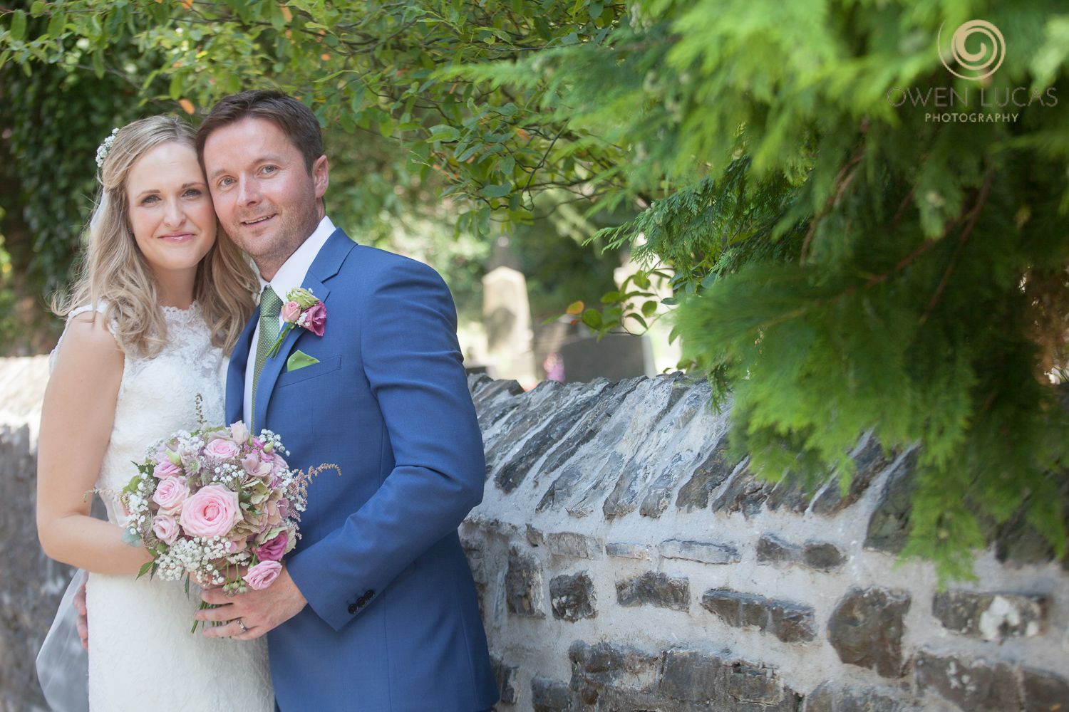 Couple poses for wedding photos in front of stone wall with lush greenery.
