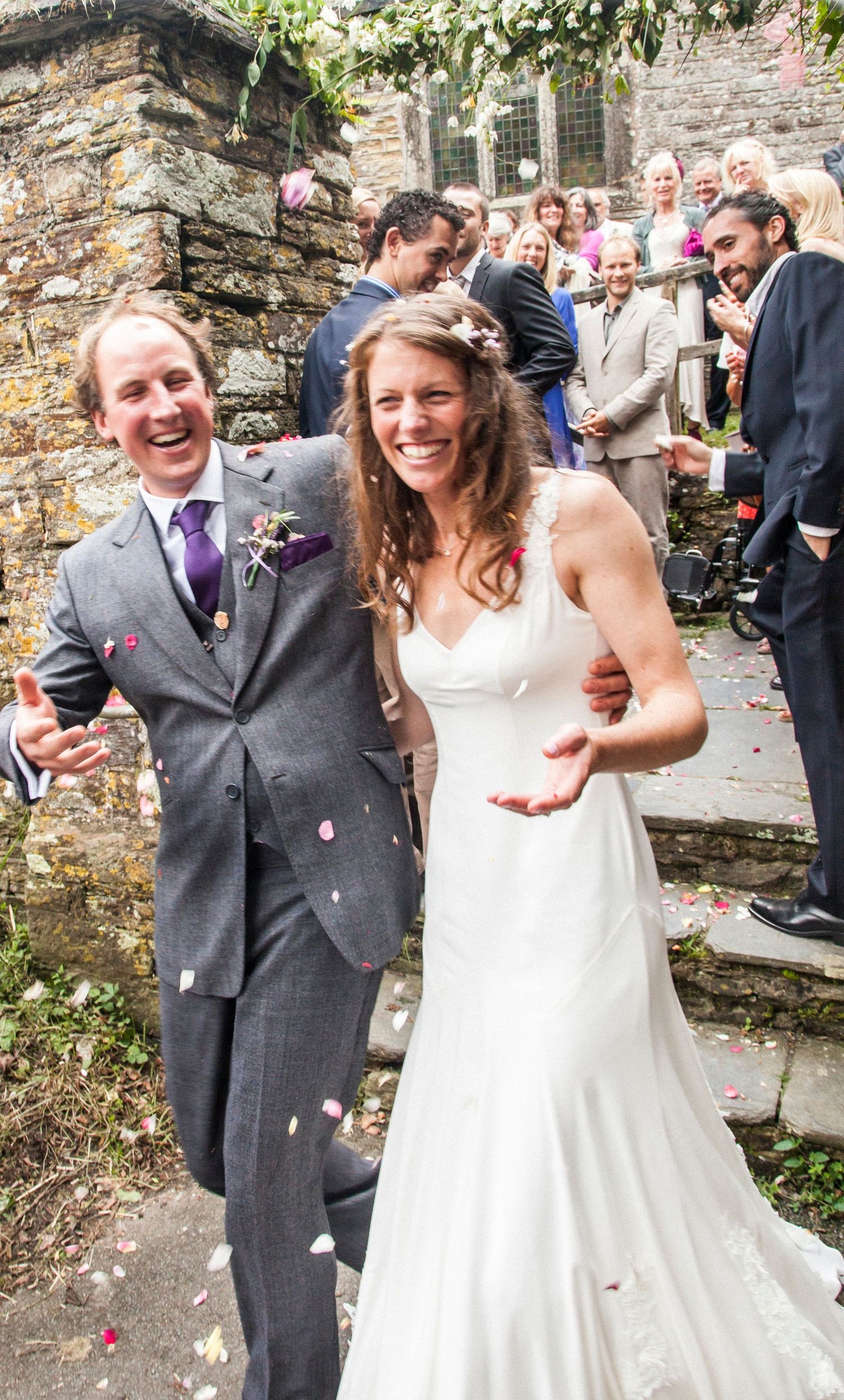 Joyful wedding couple laughing while confetti falls on stone church steps.