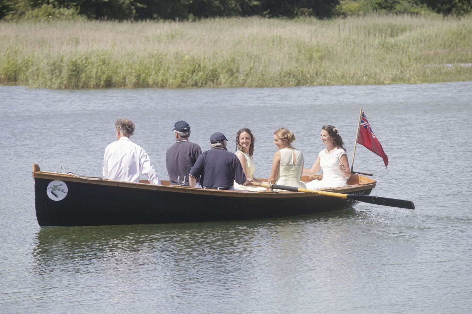 A wooden boat carrying passengers glides across a calm lake with tall grass along the shoreline.