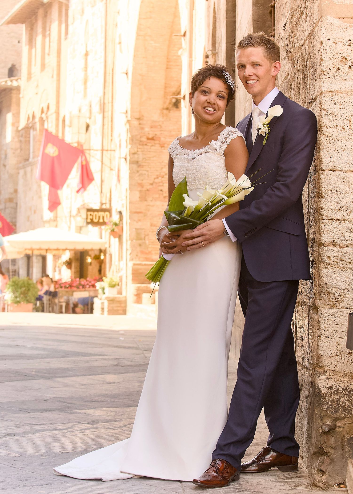 Bride and groom posing on a sunlit Italian cobblestone street, bride holding white calla lilies bouquet in elegant wedding attire.