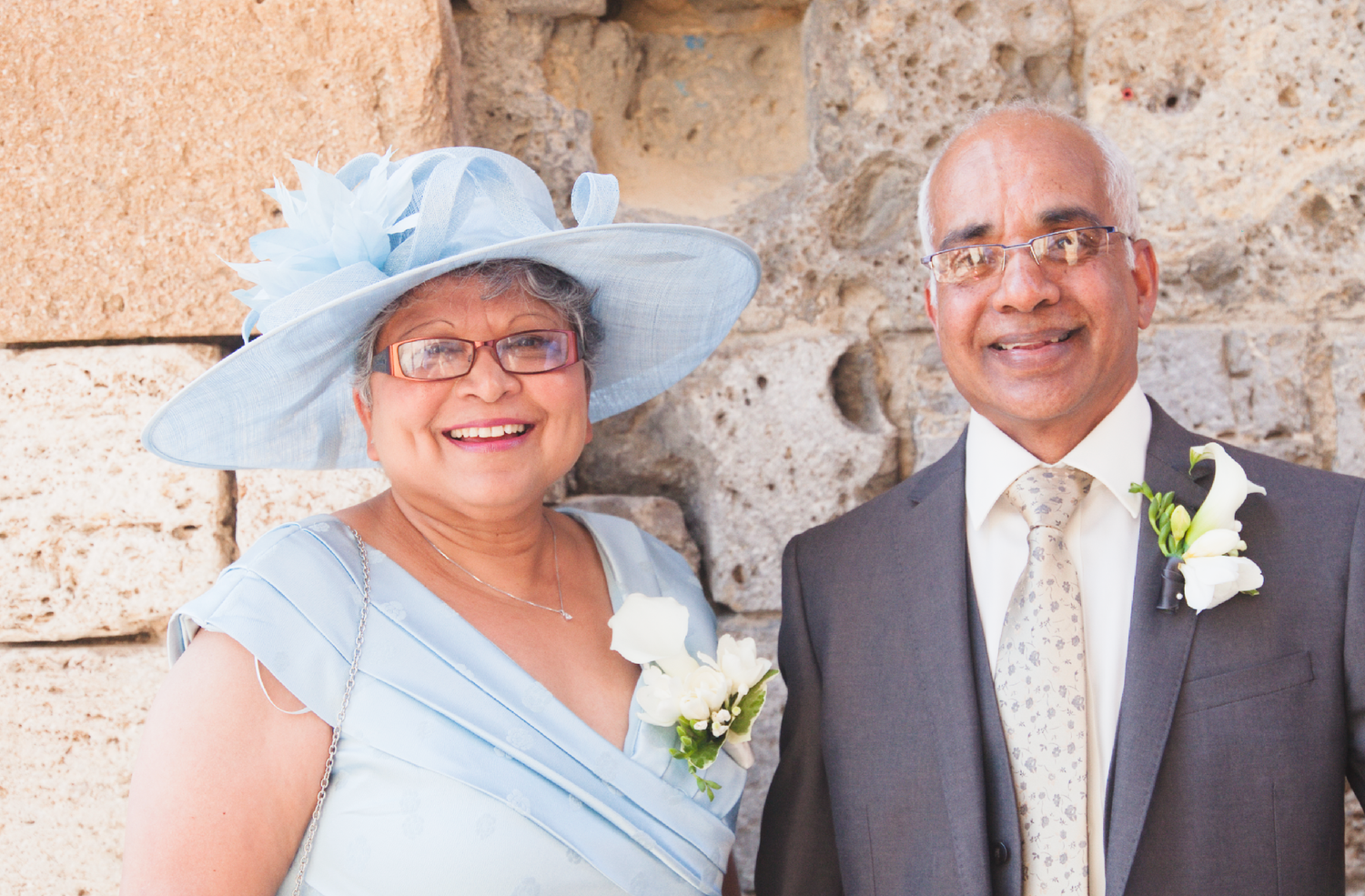 A couple in formal attire poses against a stone wall at an elegant outdoor event.