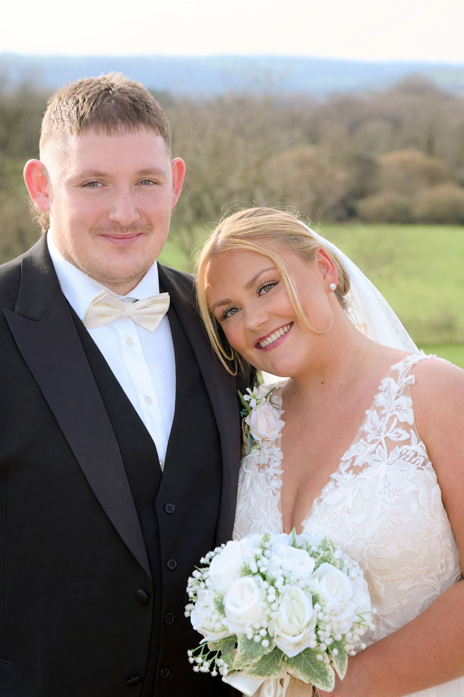 Newlyweds pose for romantic outdoor portraits with white bouquet against scenic countryside.