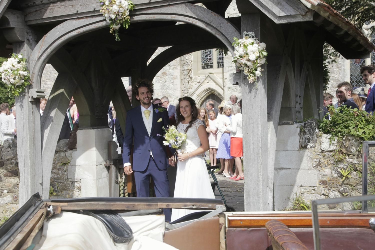 Wedding party gathering under a stone church archway decorated with white flowers.