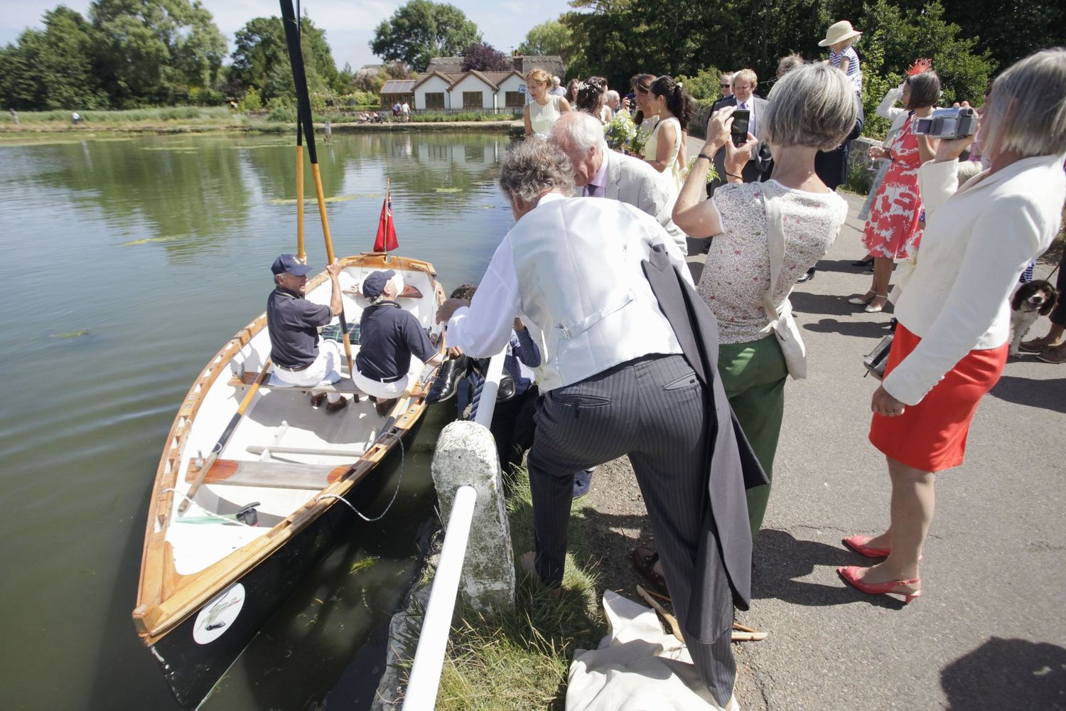 Onlookers assist passengers boarding a wooden boat from a lakeside path.