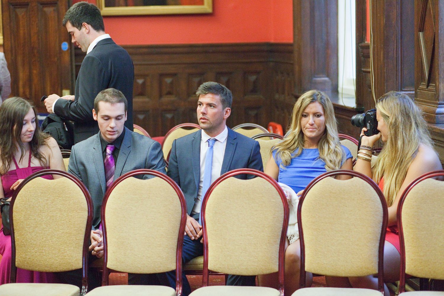 Wedding guests seated in formal chairs in a room with red walls and wooden paneling.