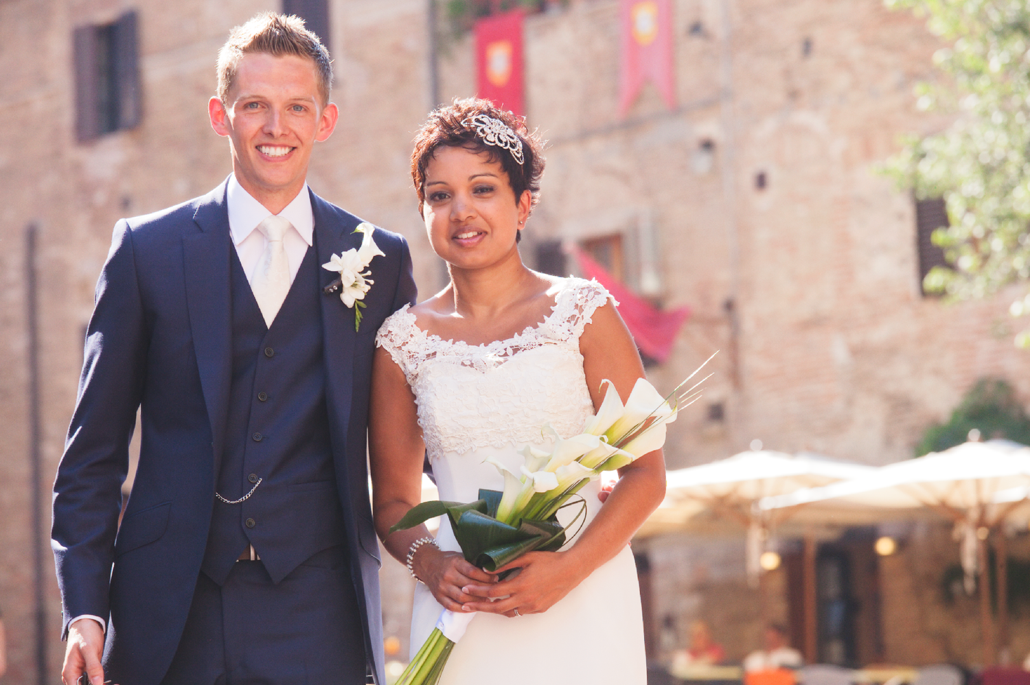 Wedding couple poses against historic stone building backdrop in sunlight.