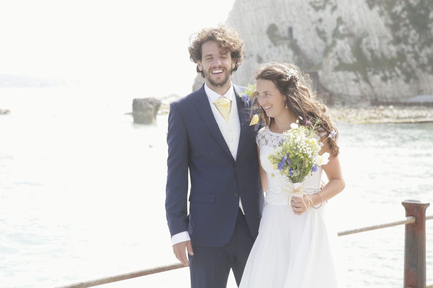 A couple in wedding attire embraces near a coastal railing with ocean views.