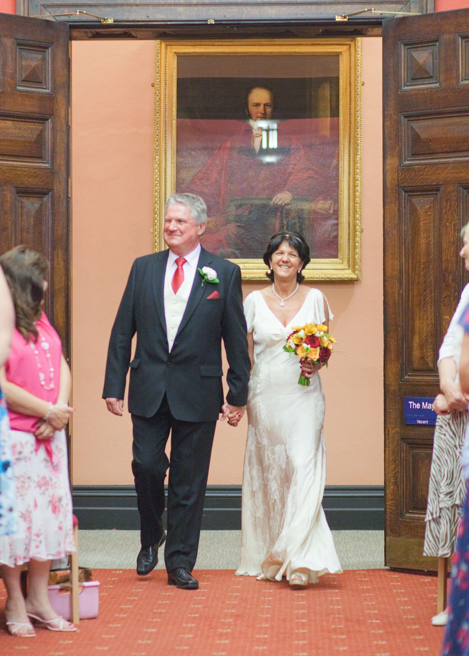 Wedding party members proceed down a hallway with coral walls and ornate wooden doors.