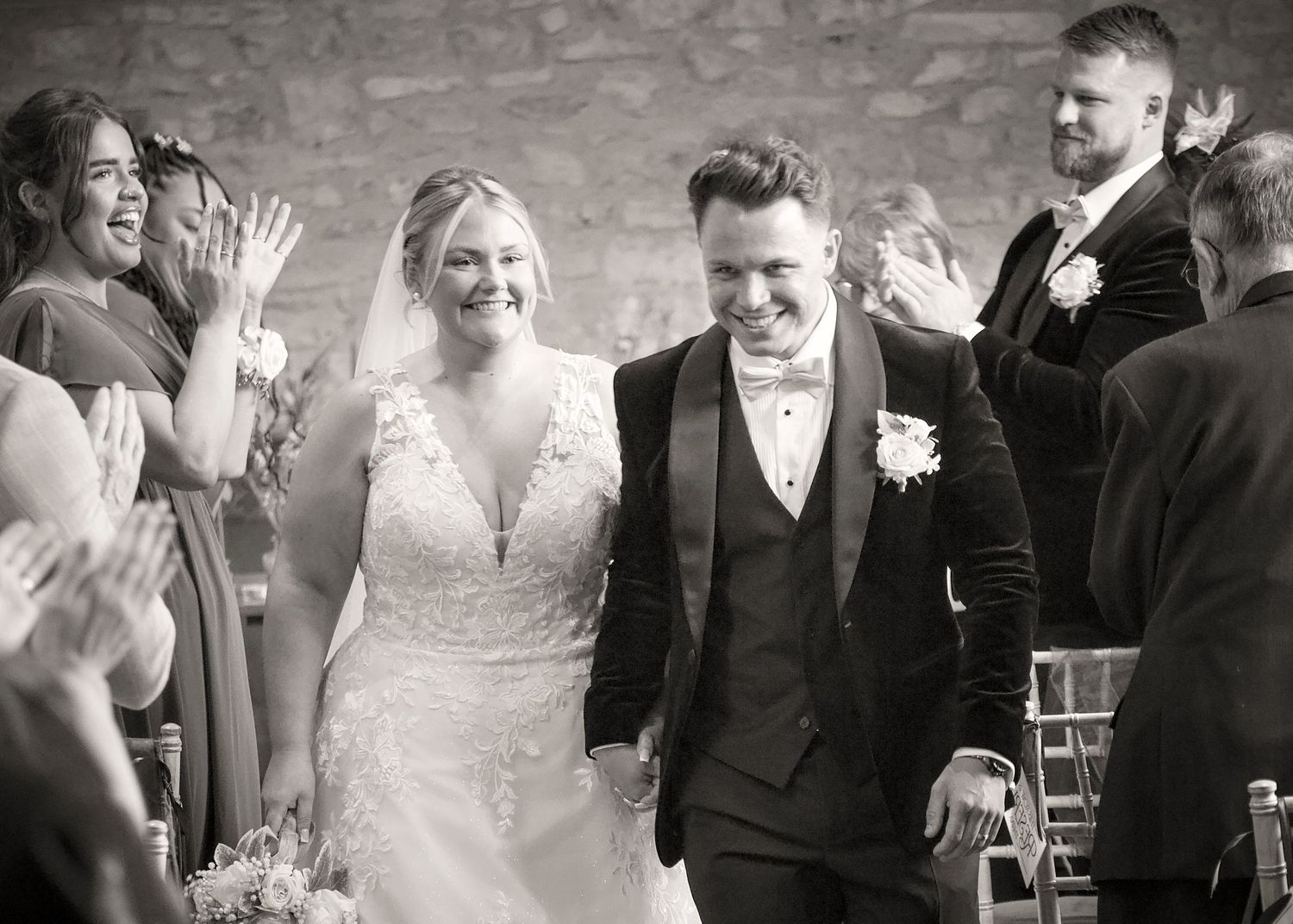 A black and white photo of newlyweds walking down the aisle while guests applaud.