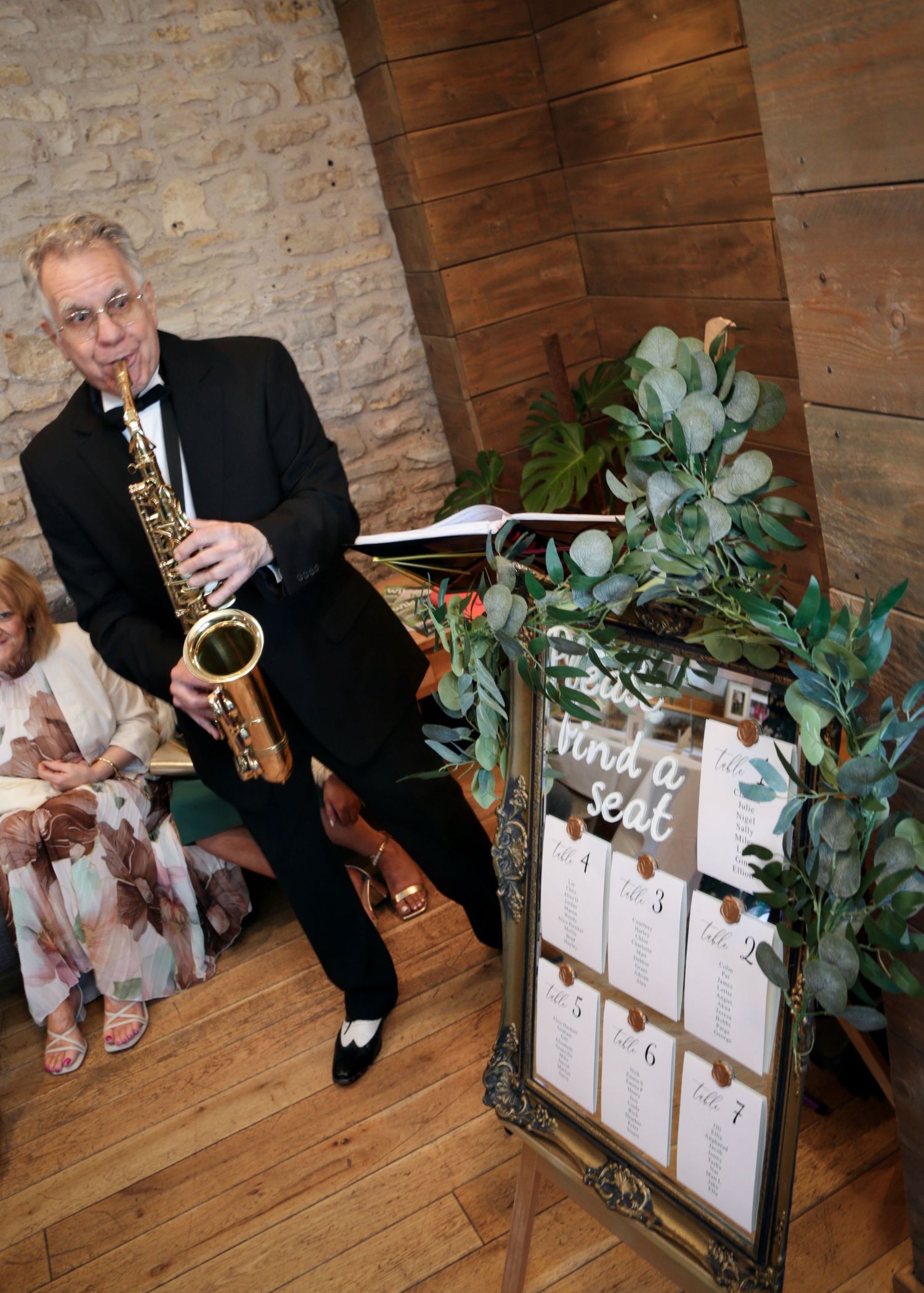Musician plays saxophone next to a decorative seating chart display at a rustic venue.