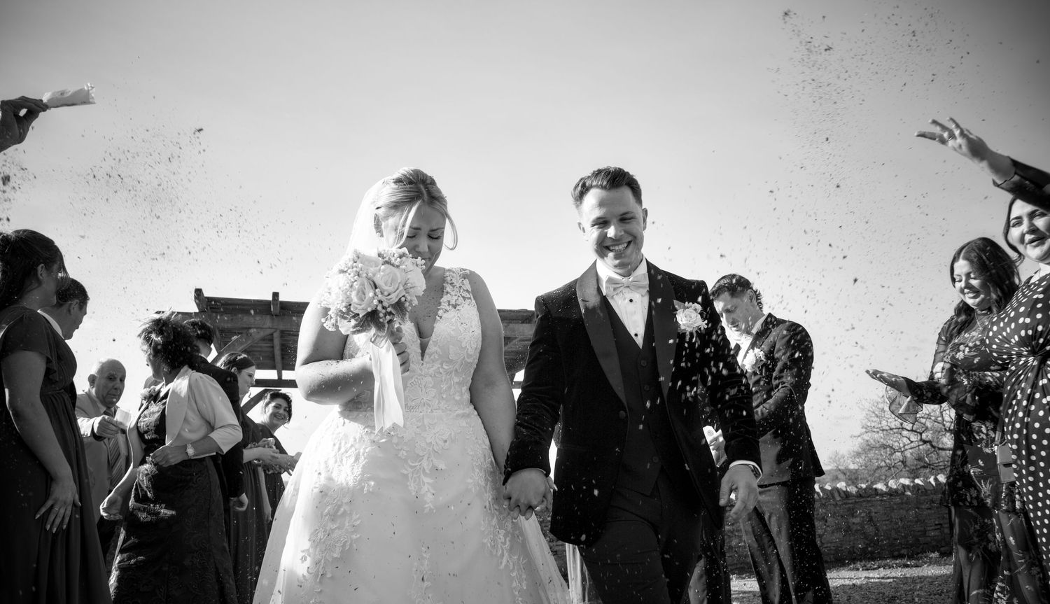 A black and white photo of newlyweds walking through confetti after their wedding ceremony.