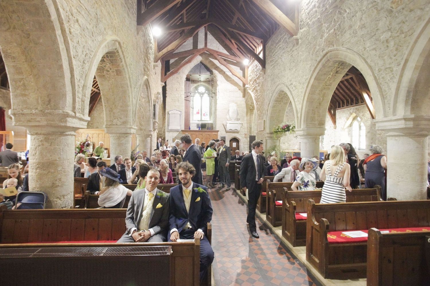 Wedding guests seated in wooden pews inside a historic stone church with vaulted arches and natural lighting.