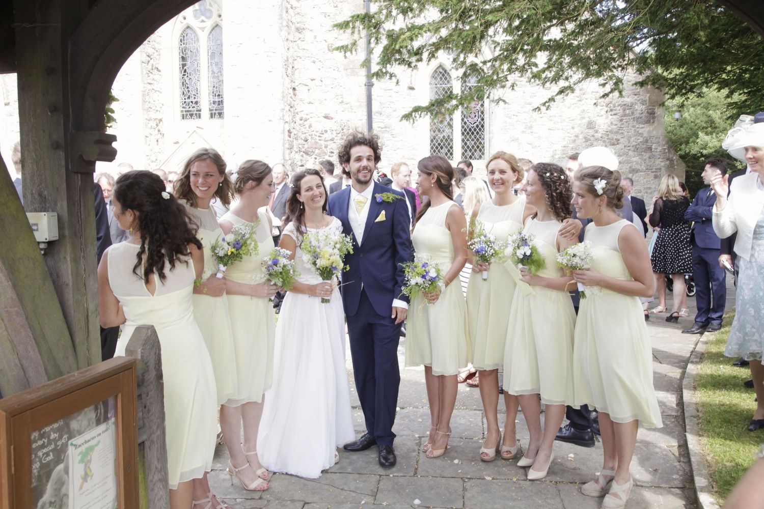 Wedding guests in pale yellow dresses and navy suits lined up outside historic stone church.