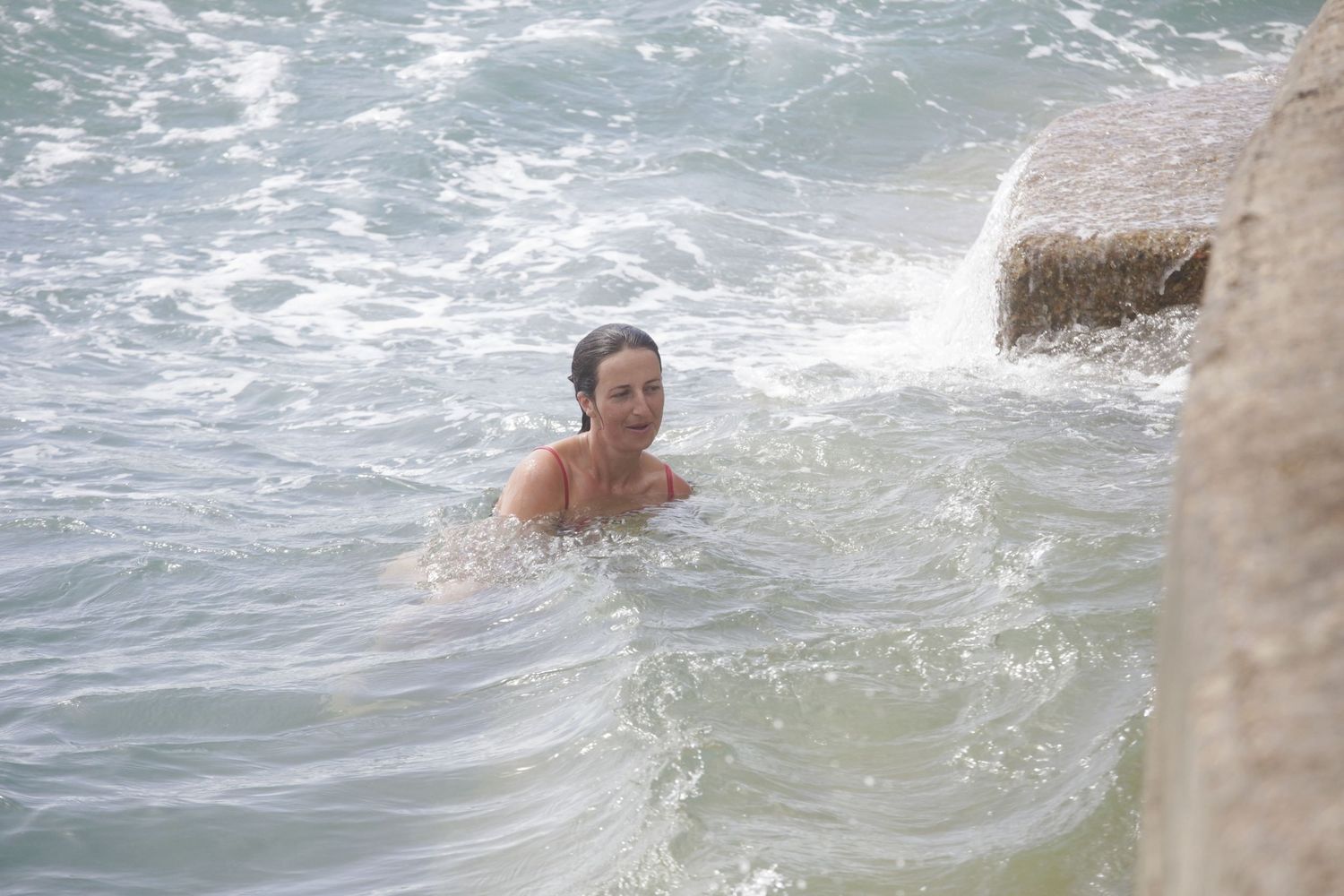 People swimming and splashing in blue ocean waters near a rocky shore.