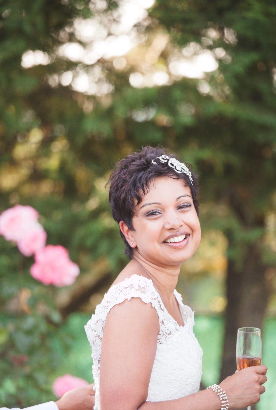 A bride wearing a lace dress and crystal hairpiece smiles while holding a champagne glass.