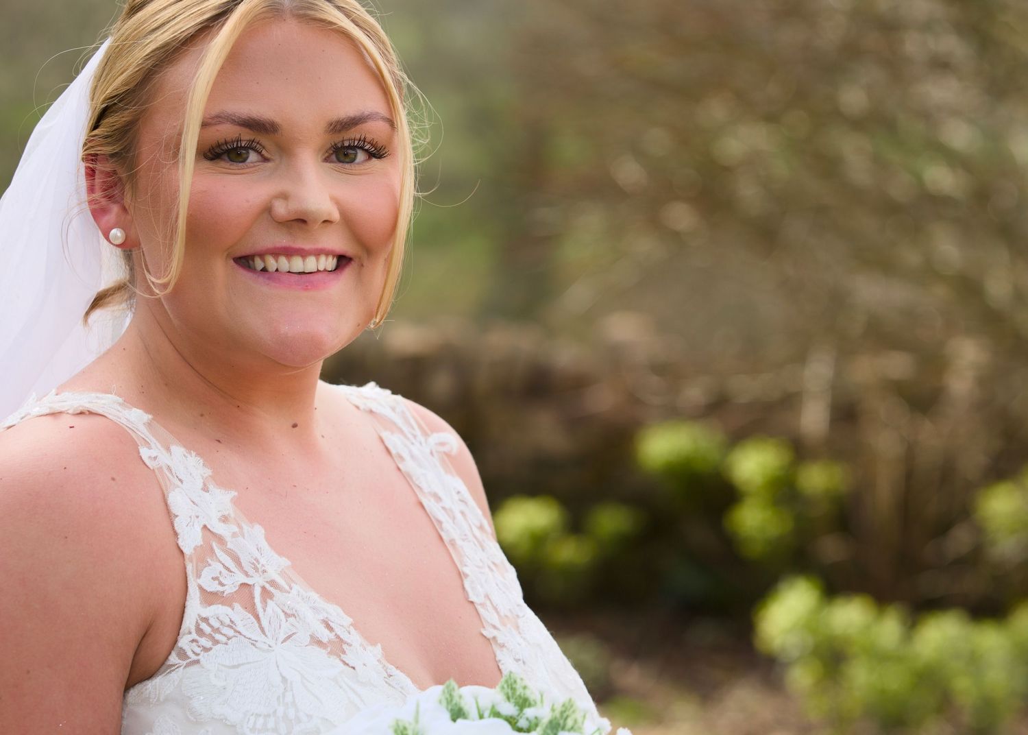 Bride in lace wedding dress smiles outdoors with natural background.