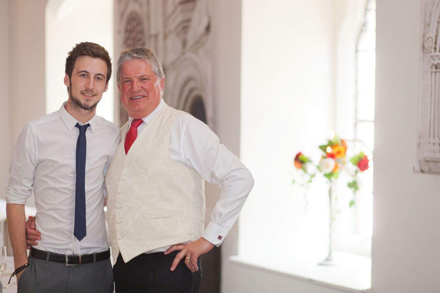 Guests in formal attire pose together in a bright, white-walled venue with windows.