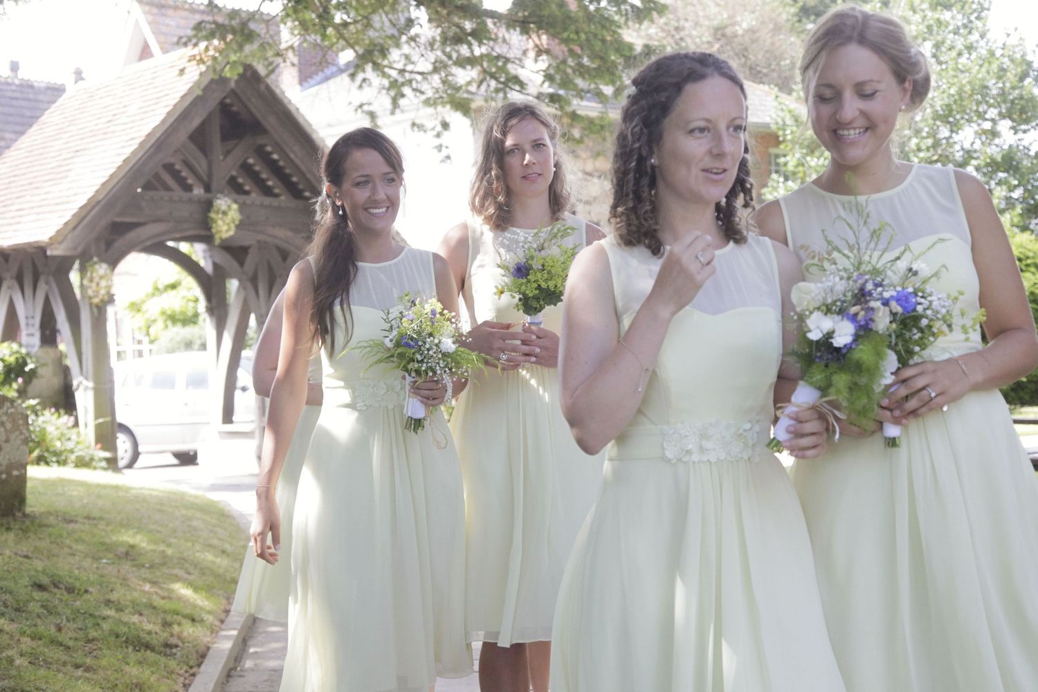 Bridesmaids in pale yellow dresses carrying bouquets outside a church entrance.