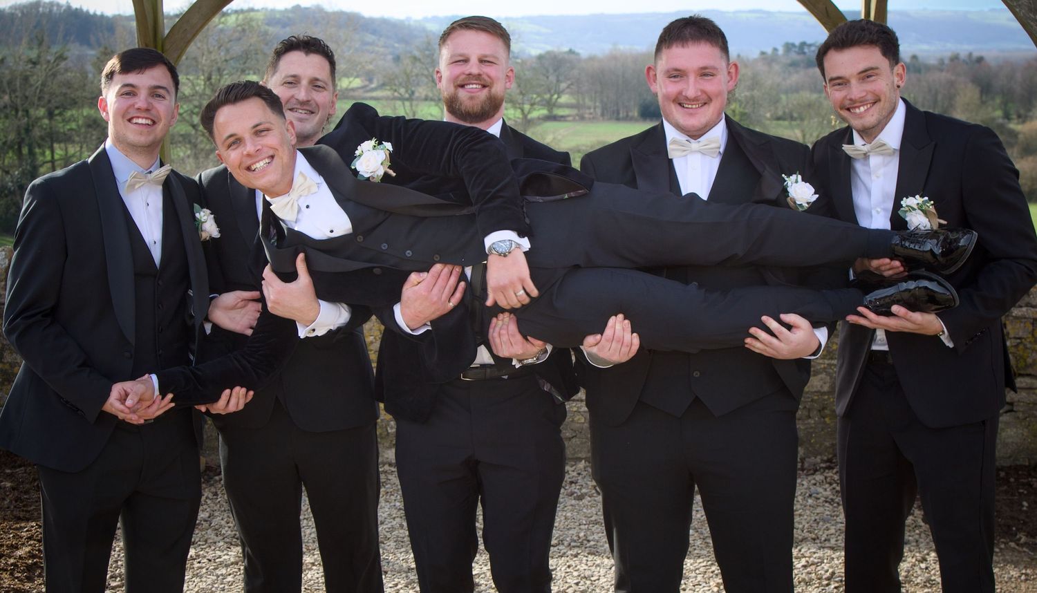 Groomsmen in black suits pose together outdoors for formal wedding photos.