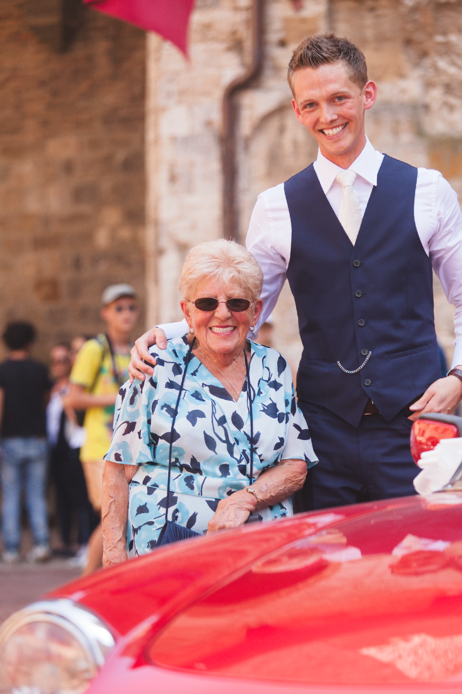 Guest in blue floral dress seated next to vintage red car at outdoor event.