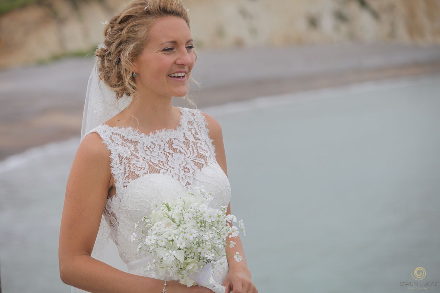 A bride in a lace wedding dress holds a white bouquet against a coastal backdrop.