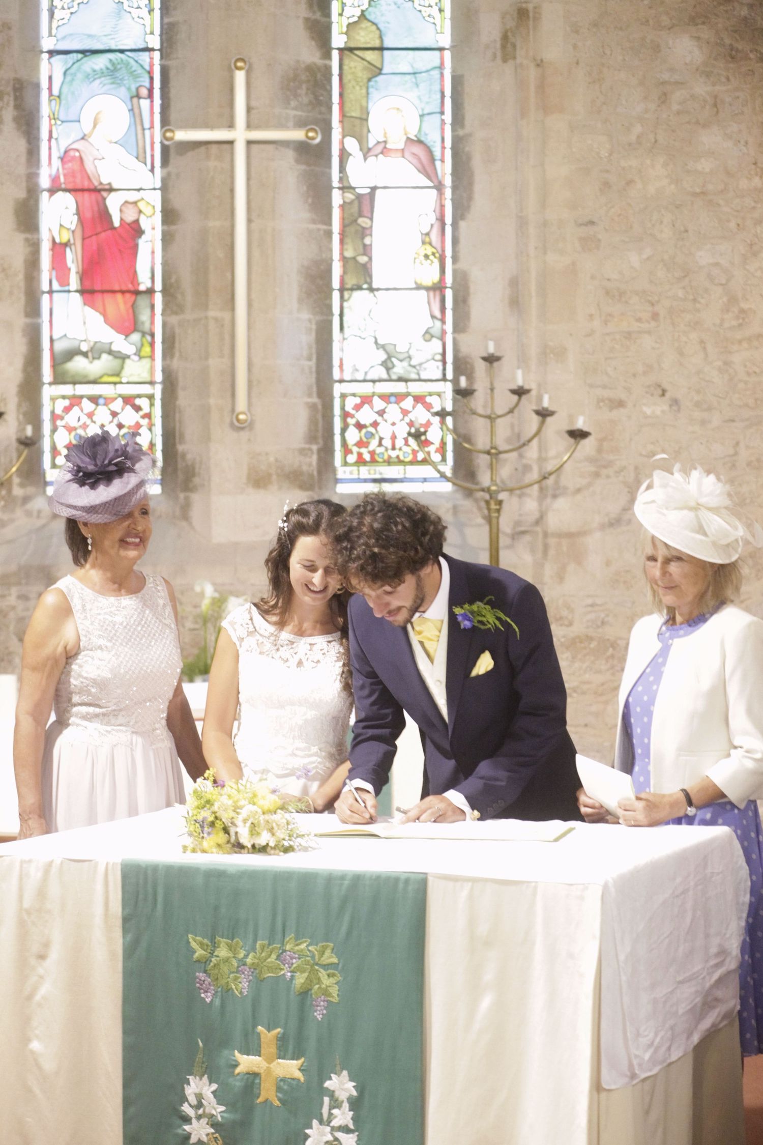 Wedding party signing documents at a table in front of stained glass windows during ceremony.