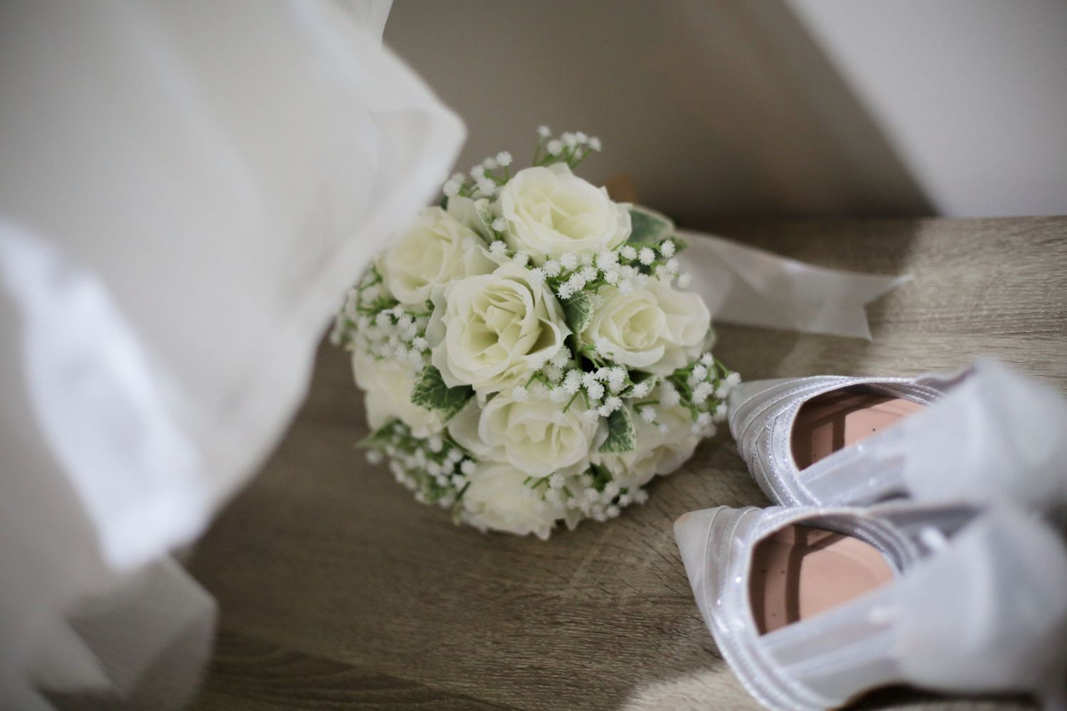 Classic white rose wedding bouquet next to bridal shoes on wooden surface.