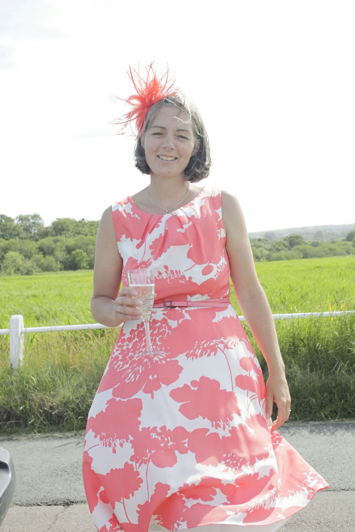 Person in coral floral print dress twirling in sunny meadow with white fence.