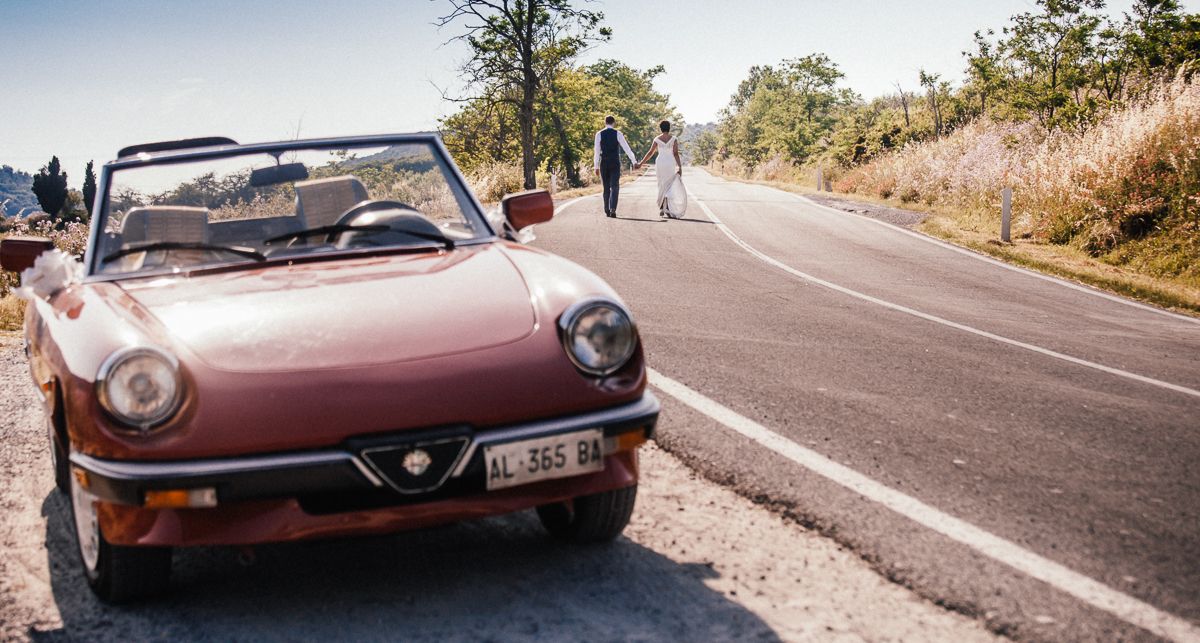 Bride and groom begin their journey as a married couple in Tuscany, Italy. They walk away from a red Triumph Spider at a destination wedding