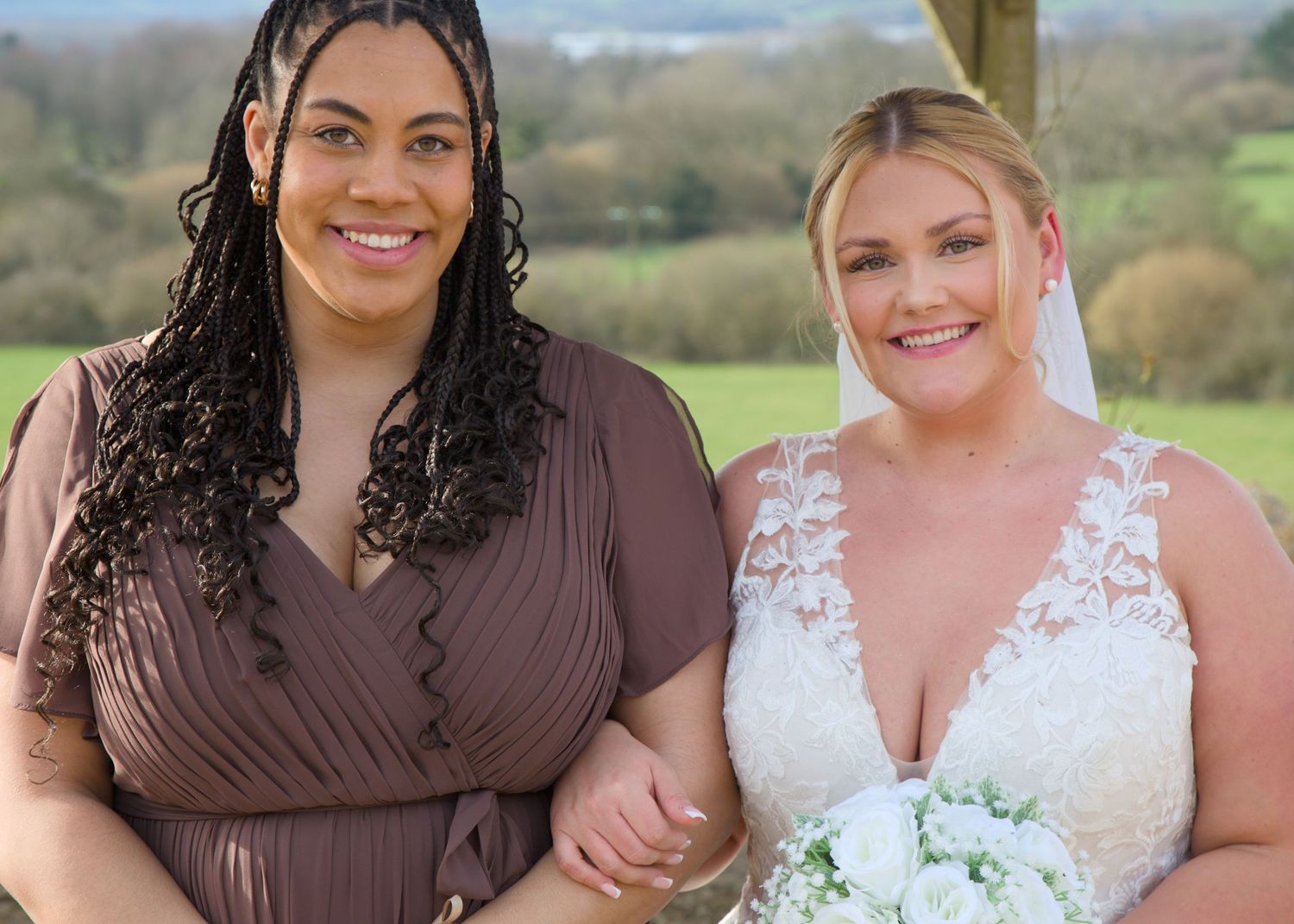 Wedding party members in brown dresses and white gown pose together outdoors with scenic countryside backdrop.