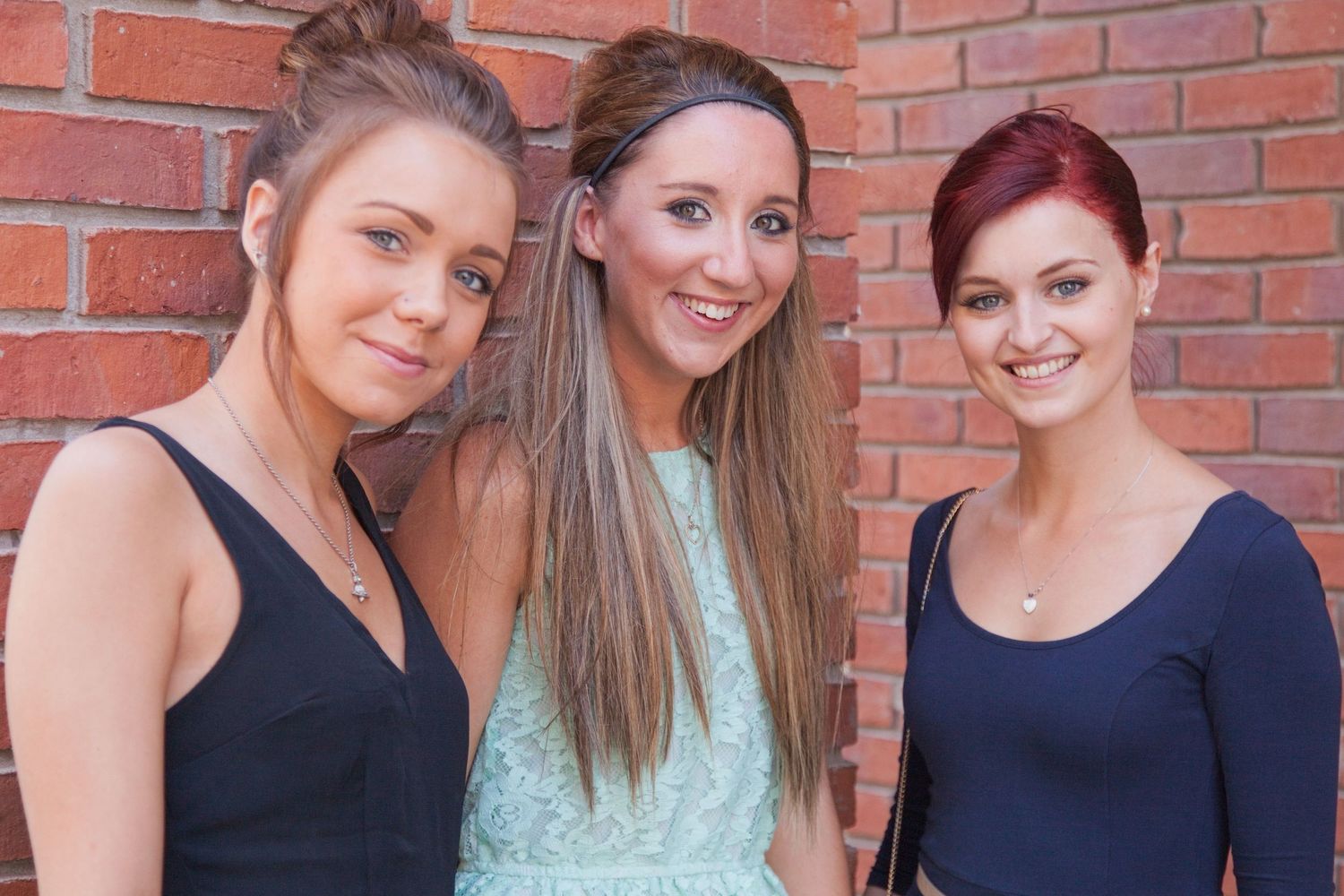 Group of women in formal dresses pose together against brick wall background.