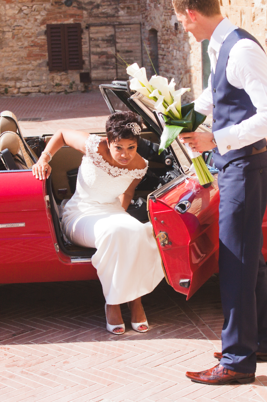 Wedding couple poses with white calla lily bouquet against rustic brick wall and red carpet.