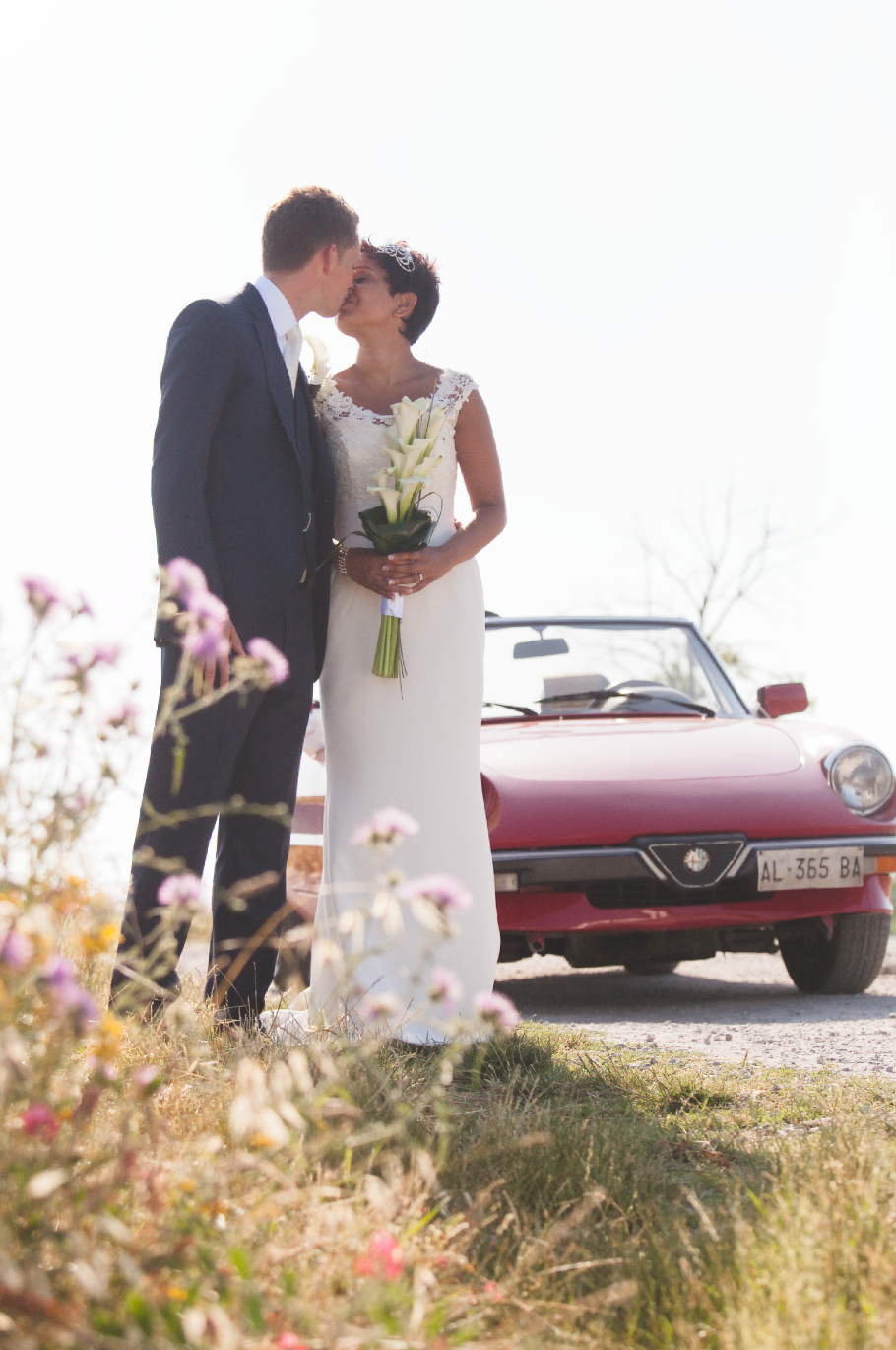 A romantic wedding photo with a classic red vintage car against a scenic countryside backdrop.