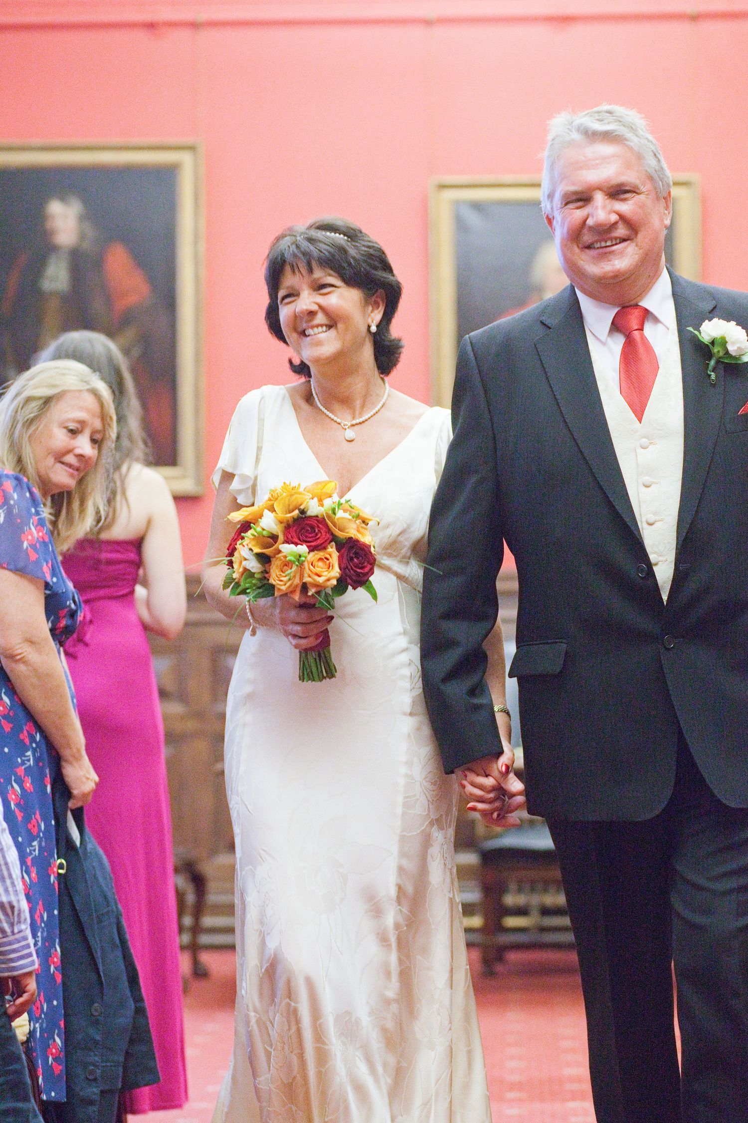A happy wedding couple standing in a room with red walls and portrait paintings.