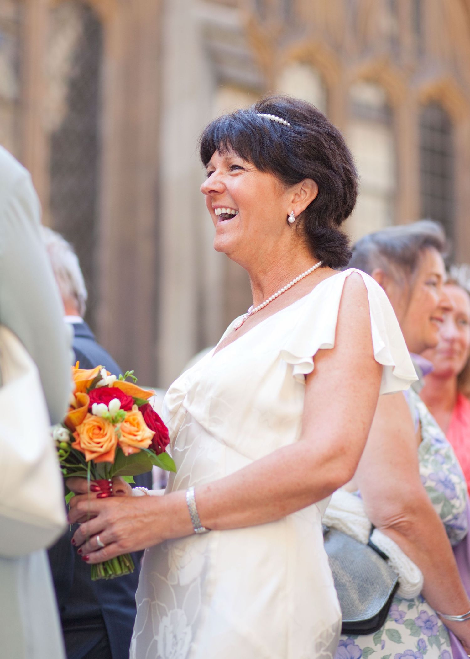 Wedding attendee in cream-colored dress laughs while holding a bright floral bouquet.