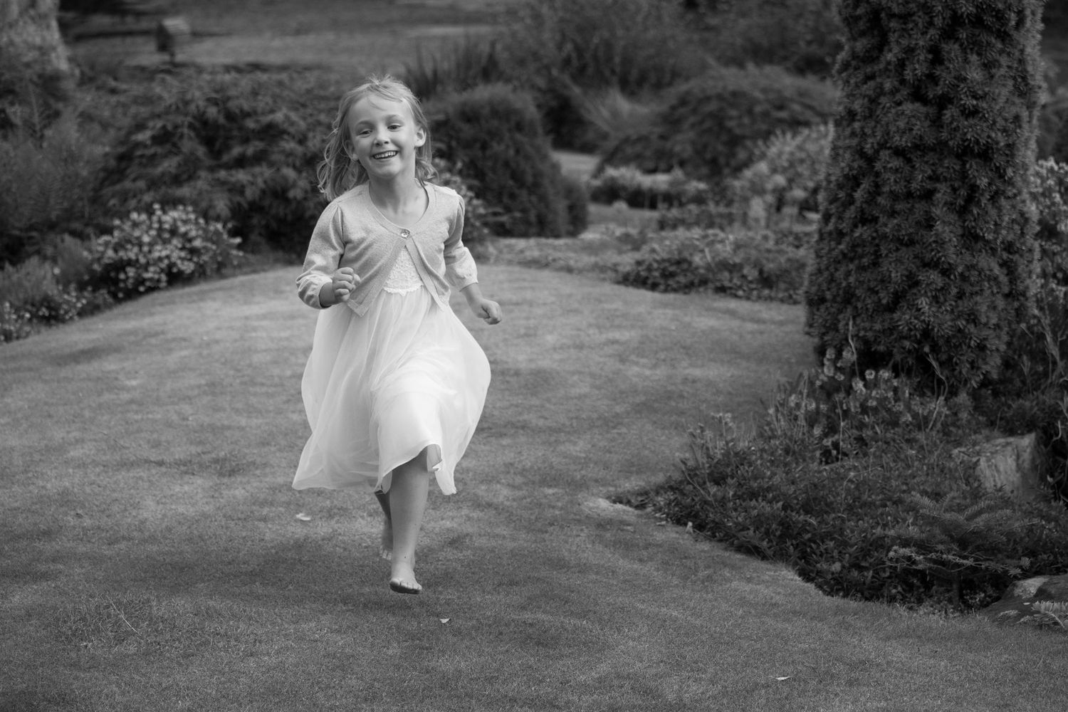 Young flower girl in white dress running joyfully through garden in black and white photo.
