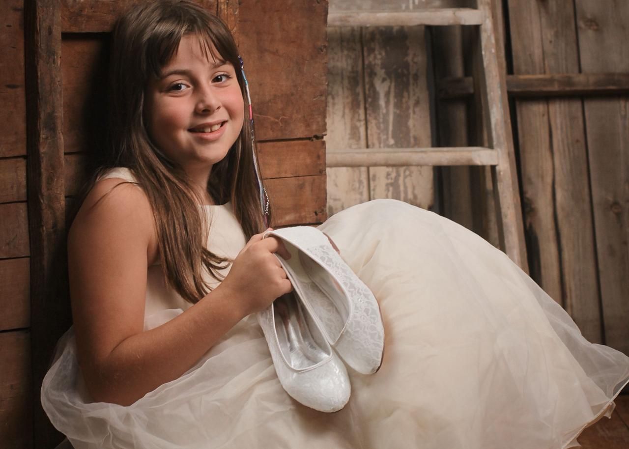 A rustic barn portrait featuring someone in a white dress holding ballet shoes with warm lighting and wooden backdrop.