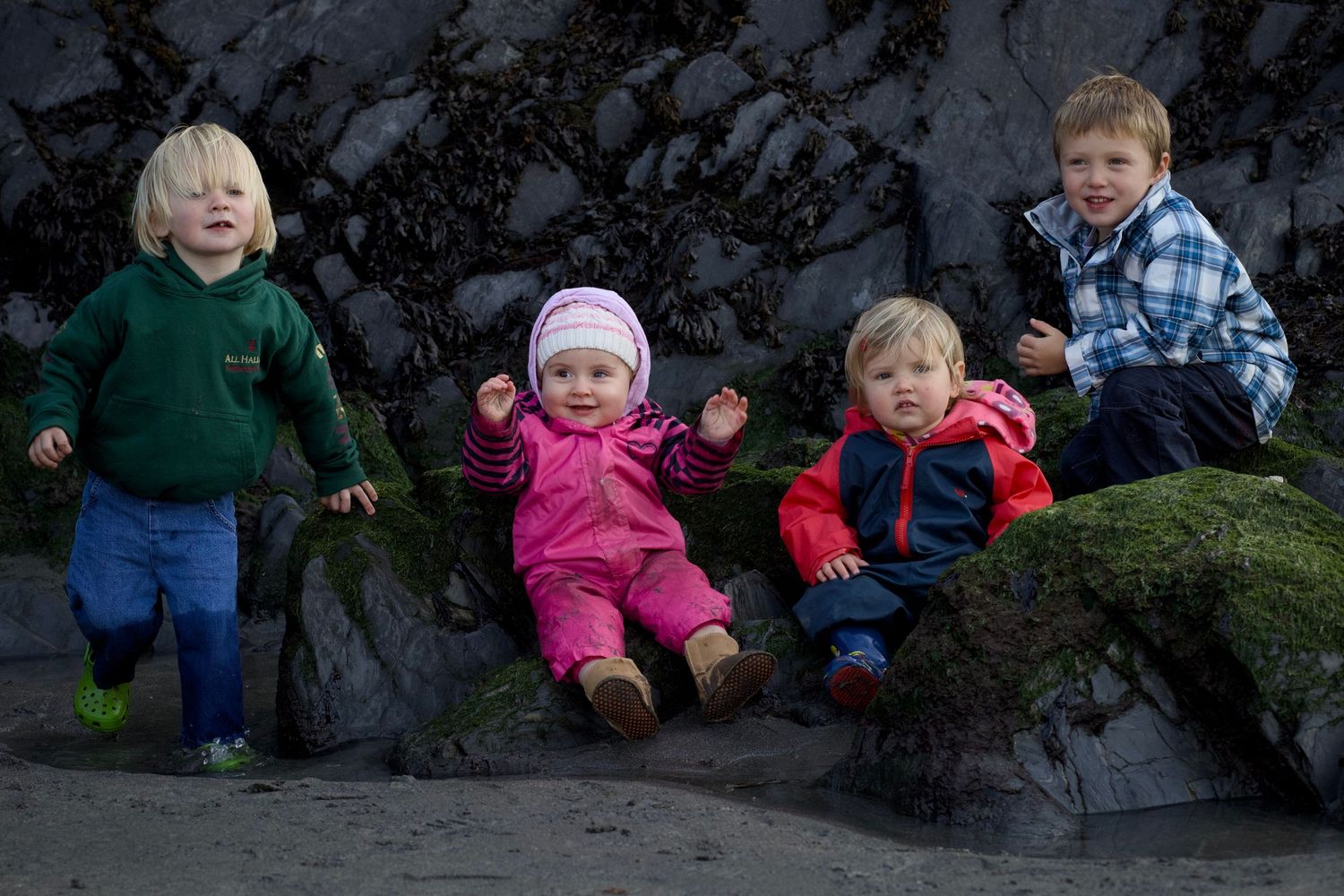 Four children sit together on a mossy rock ledge against a dark stone wall during winter.