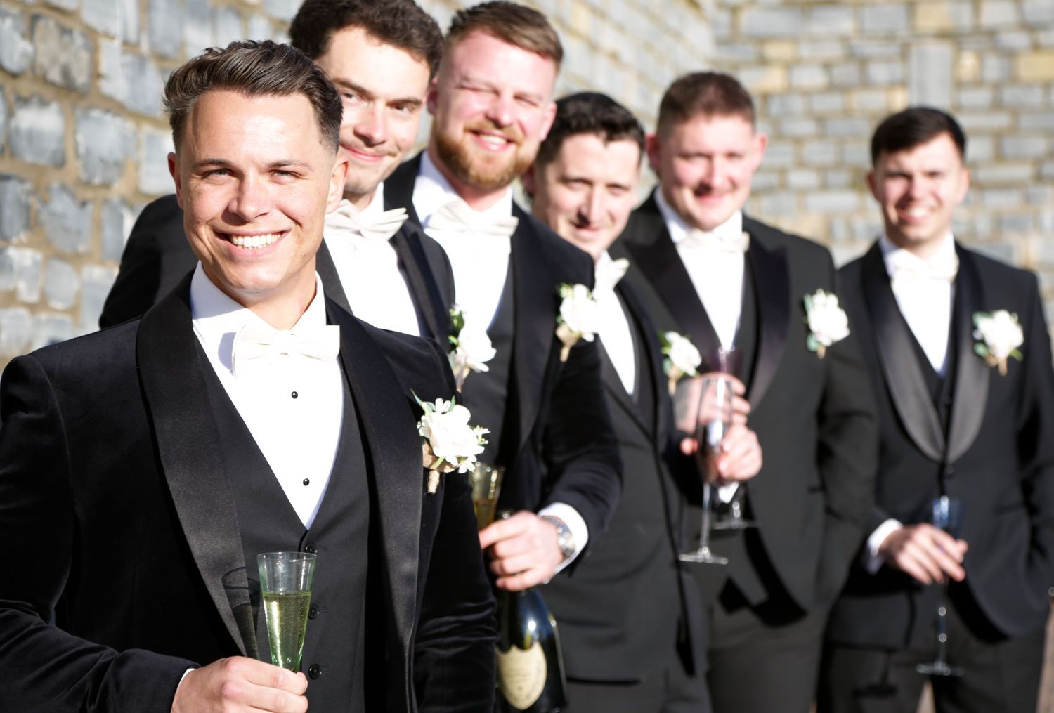 Groomsmen in formal attire pose together while holding champagne glasses outdoors.