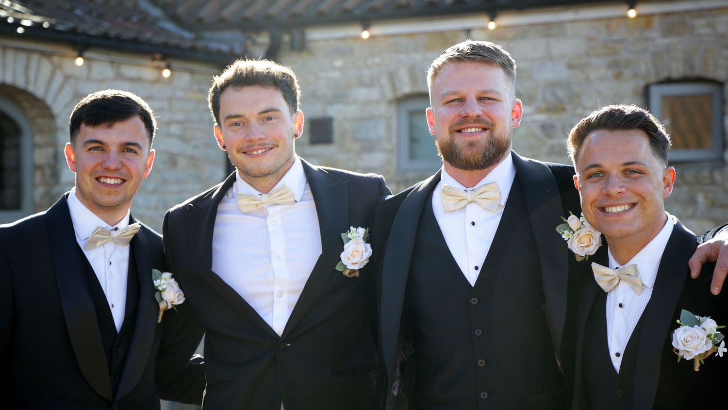 Four groomsmen in formal attire pose together outdoors at a wedding venue.
