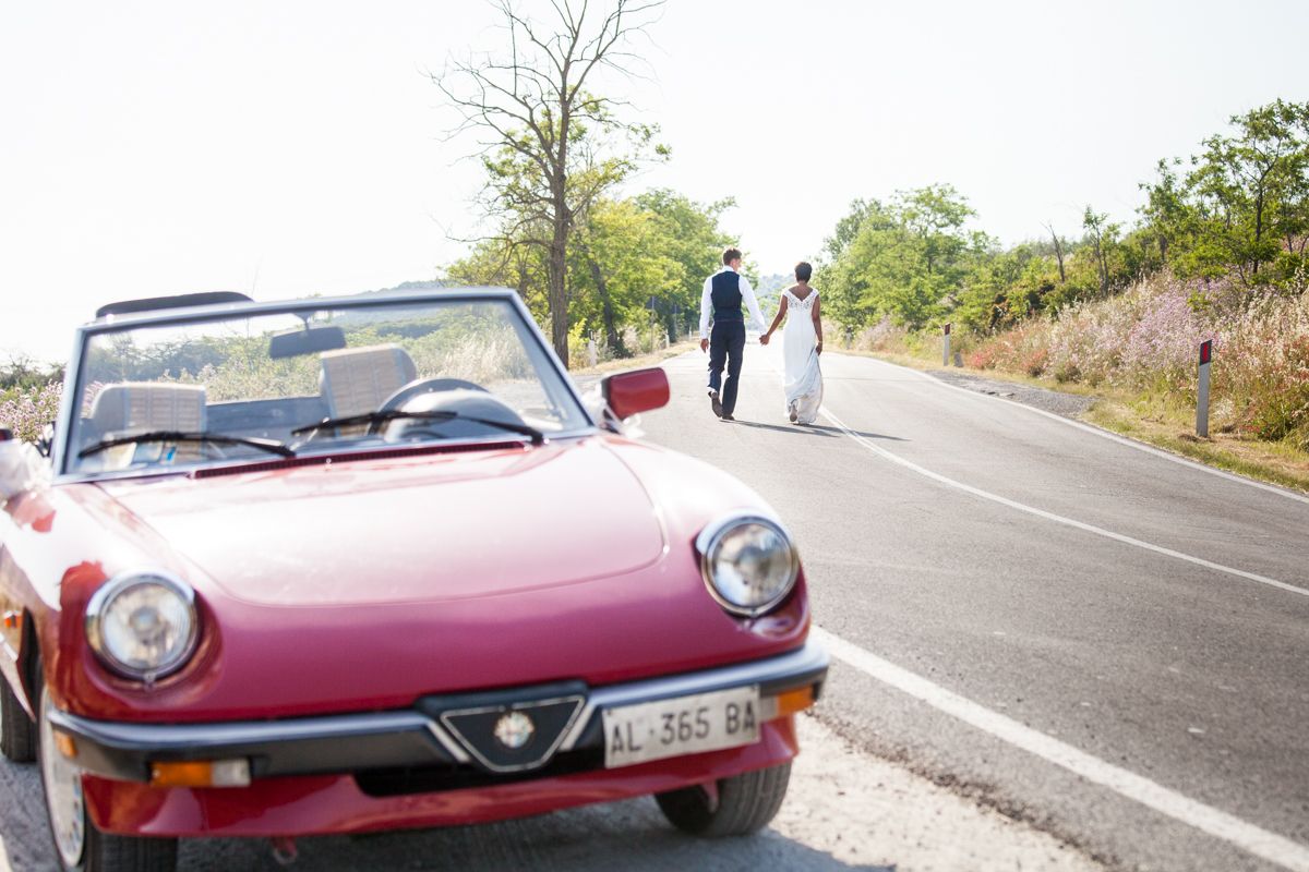 A classic red Alfa Romeo Spider convertible sports car parked on a scenic mountain road with trees in the background.