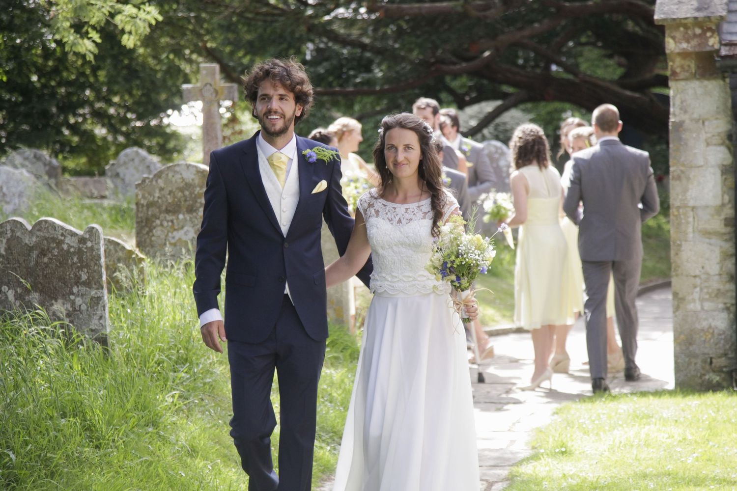 Newlyweds walking through sunny church garden path after ceremony.