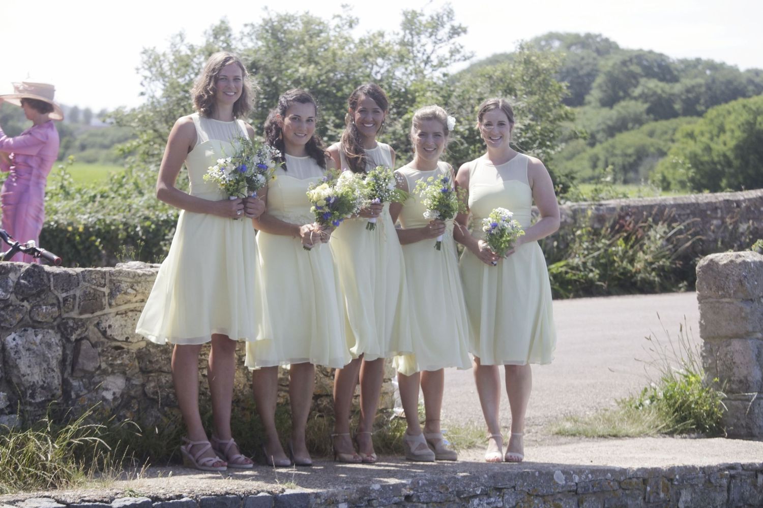 Row of bridesmaids in light yellow dresses stand along a stone wall pathway.