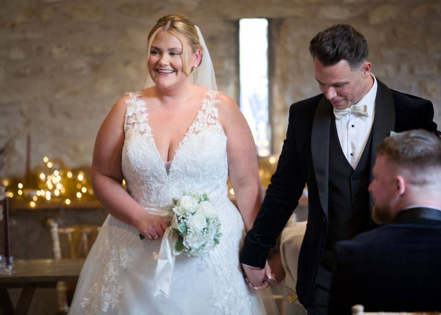 Newlyweds in formal attire proceed through their wedding ceremony in a rustic venue.