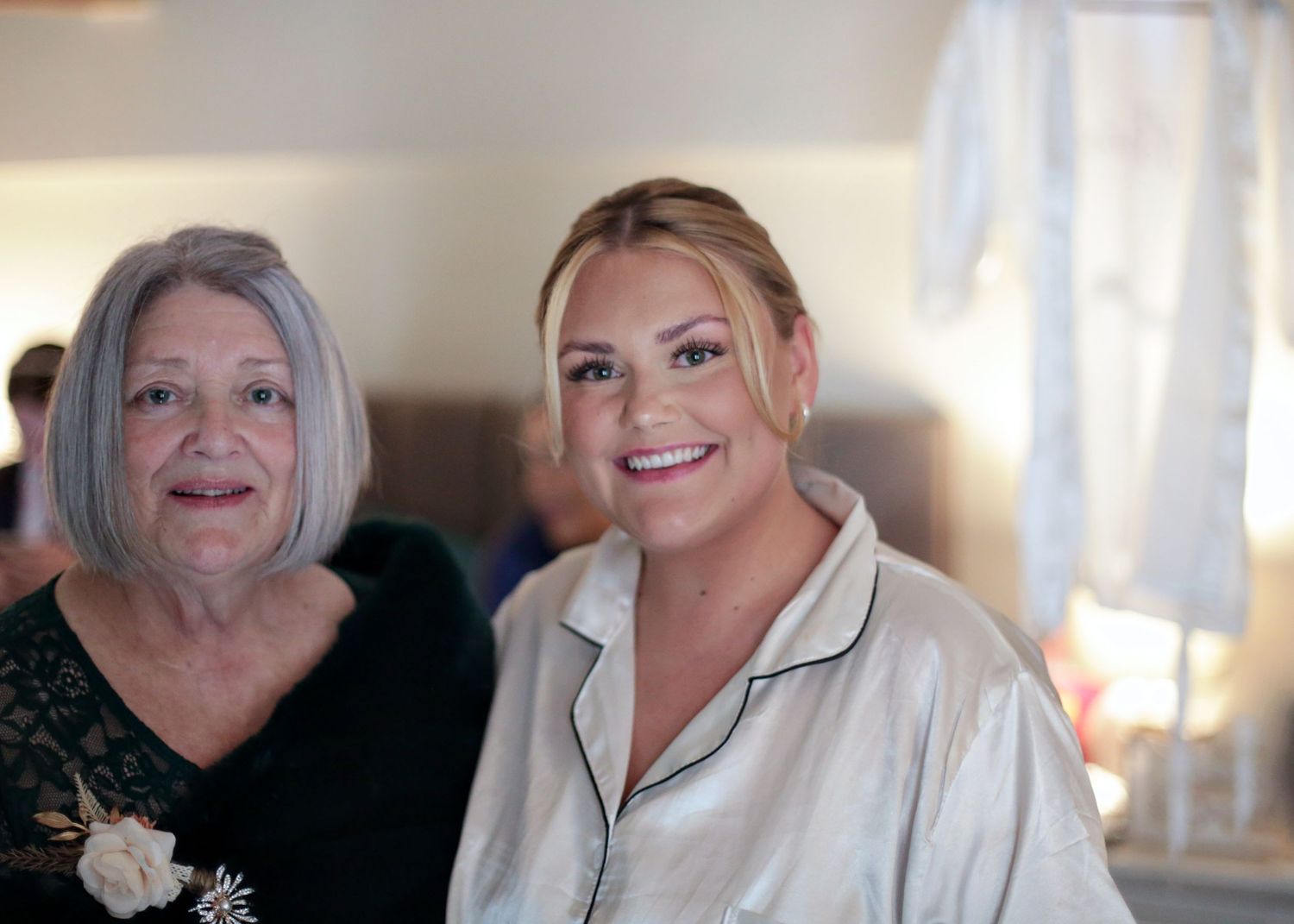 Two women smile together during wedding preparations, one in silk pajamas and one in black.
