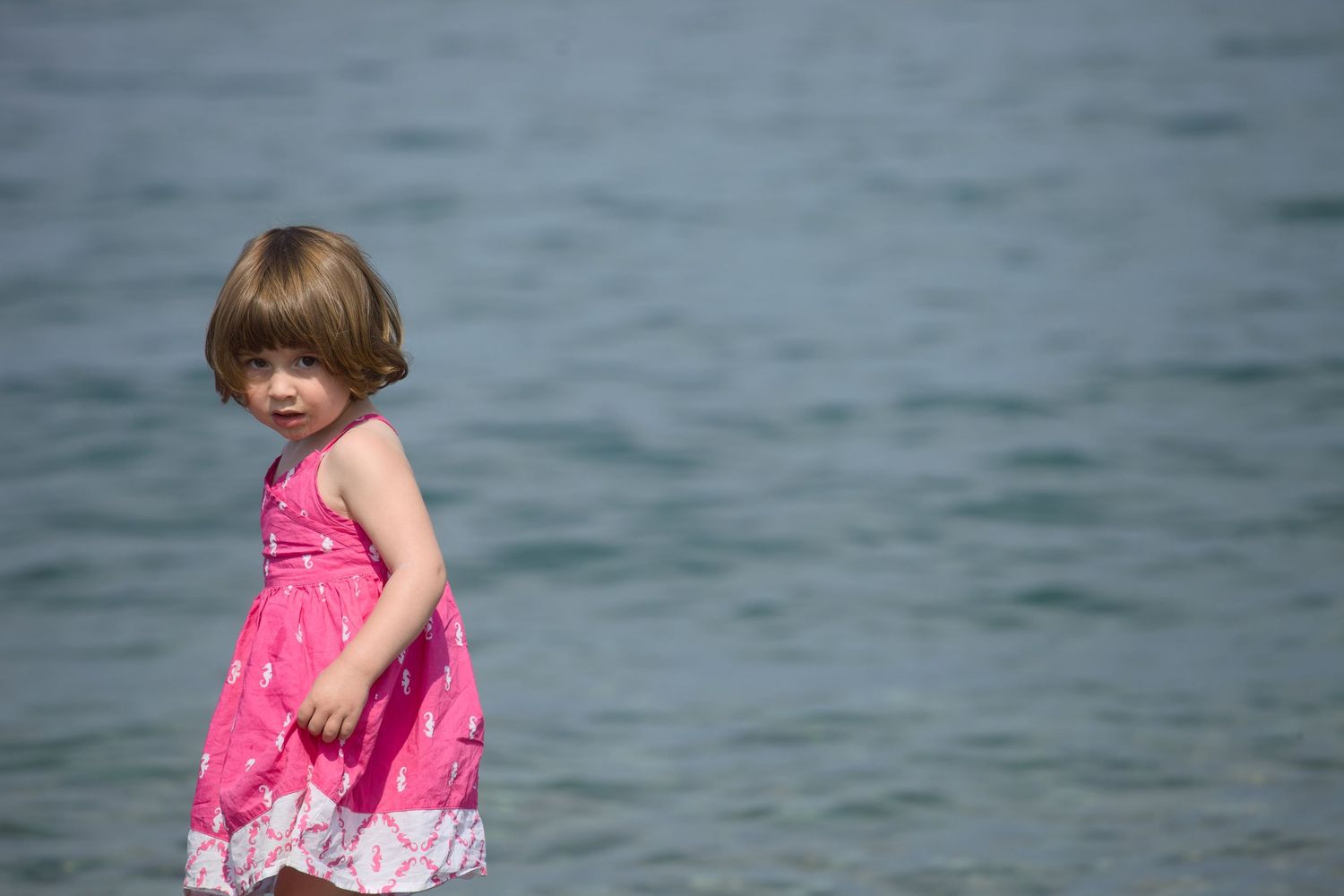 Child in a pink summer dress stands by the water on a sunny day.