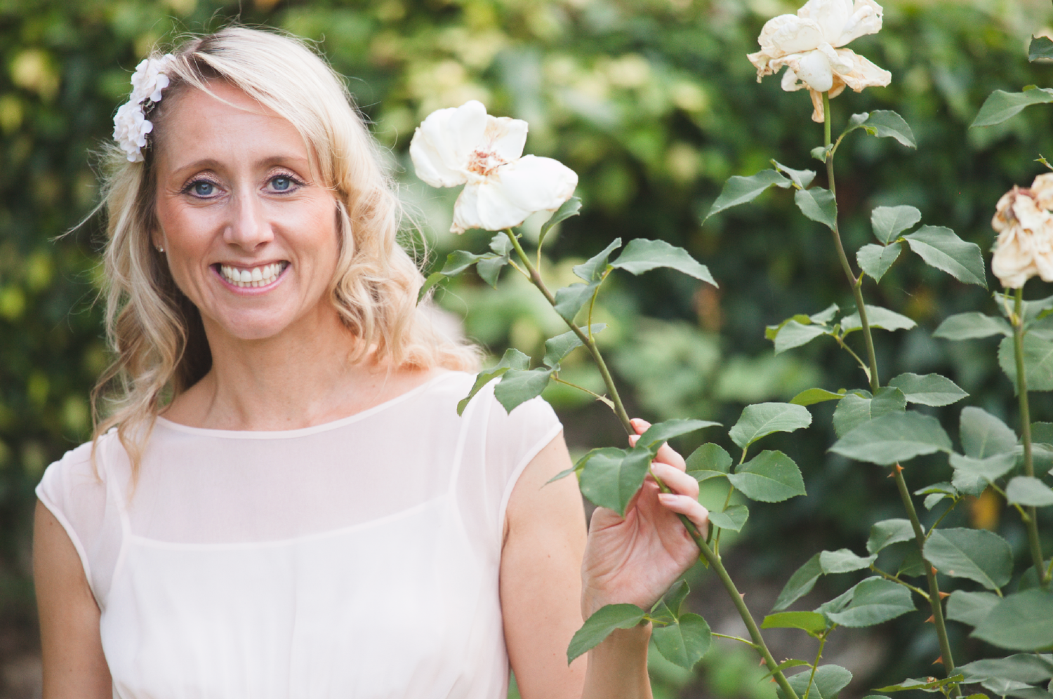 A bride in white stands near blooming roses in a garden setting.